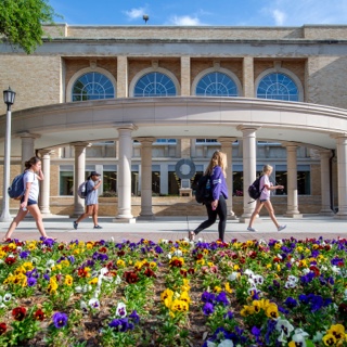 TCU students walk by a flowerbed of multi-color pansies on a sunny day near the library