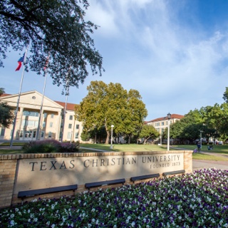 A brick sign bearing the name Texas Christian University sits behind a flowerbed of purple pansies